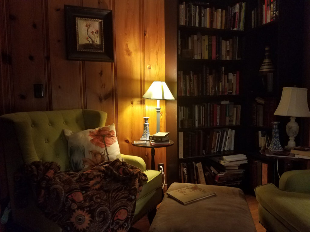 Image of a small library with pinewood paneling. Two green arm chairs sit in the corner, with small lamps and tables. One lamp is lit. There is a botanical painting on the wall, and a orange-patterned blanket on the chair.