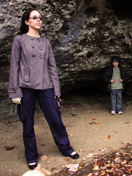 A woman in a brown-gray coat standing on a walkway under a rock outcropping, holding a dog leash. A small child is standing in the background.