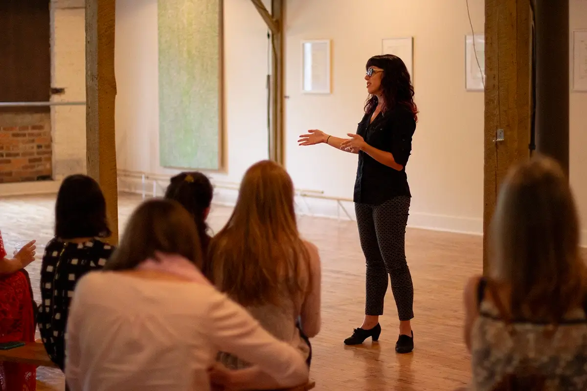 An image of Audra leading a workshop at the abrasiveMedia location in Houston Station, in 2015. Audra is wearing a black top, black patterned pants, and black shoes. She is talking with her hands as she addresses a group of people we see from the back. An abstract painting by artist Jacob Gregory is in the background.