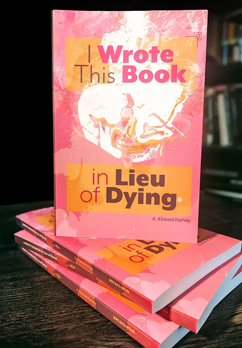 A stack of copies of "I Wrote This Book in Lieu of Dying" on a dark wood table, with a blurred bookshelf in the background.